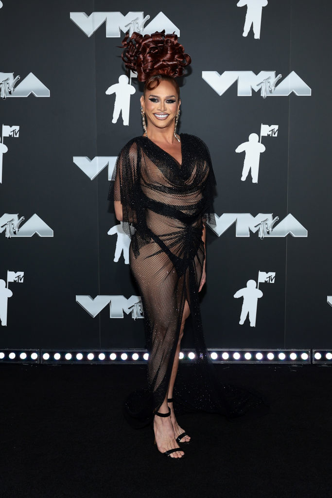 Sasha stands on the VMAs red carpet in a sheer, draped gown and high heels, with an intricate updo hairstyle