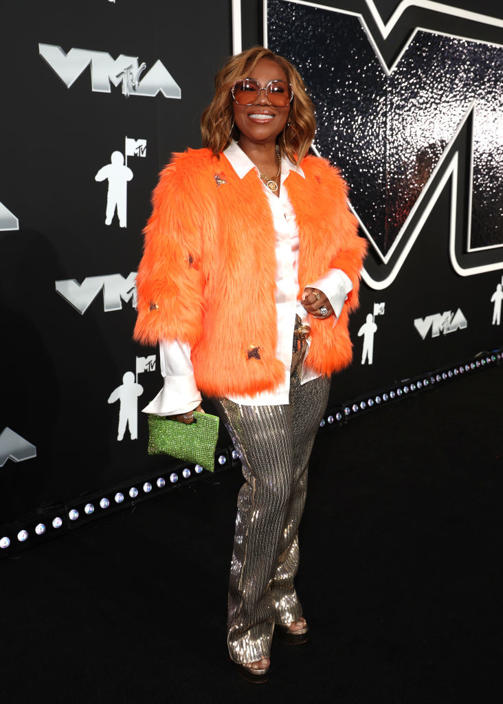 Mona at the VMAs wearing a vibrant fur coat, blouse, sparkling pants, and sunglasses, holding a purse