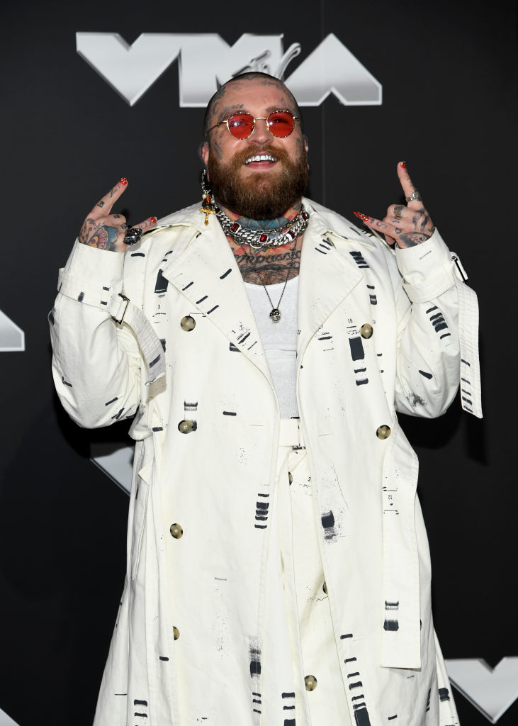 Teddy on the red carpet in a patterned trench coat and pants, wearing accessories and flashing hand signs