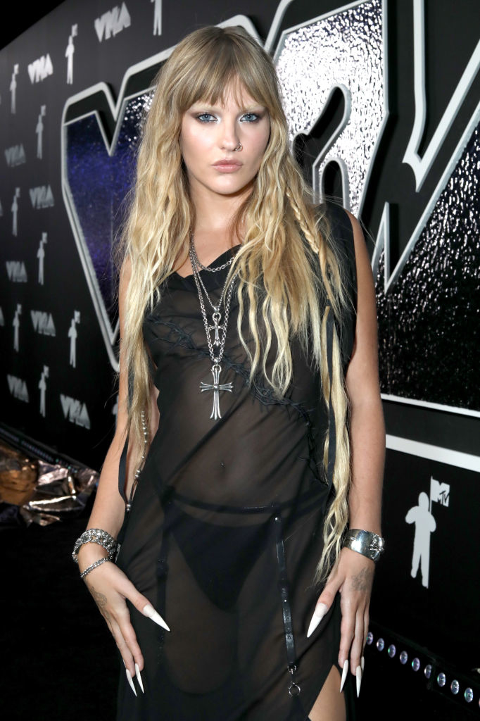 Victoria at the MTV VMAs, wearing a sheer dress with long hair and striking nails, accessorized with silver jewelry and a cross necklace