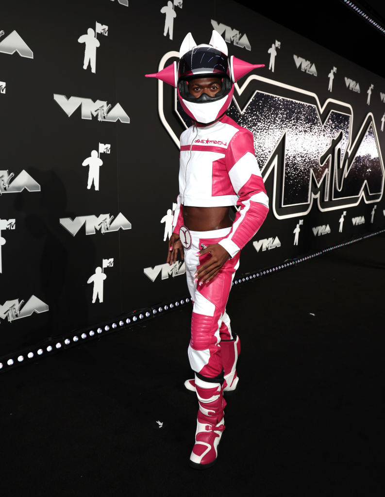 Lil Nas X in futuristic motorcycle gear on the VMAs black carpet, wearing a helmet with pink accents and matching outfit