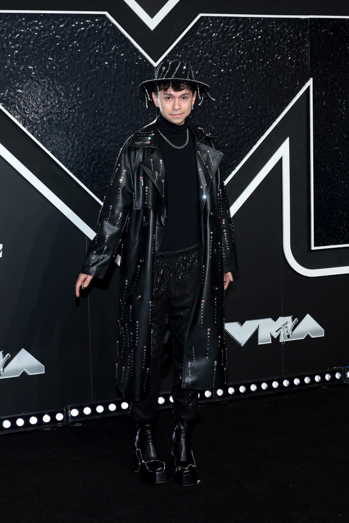 Mikey Angelo on the red carpet at a VMAs event, wearing a long leather coat, turtleneck, patterned pants, and a wide-brim hat