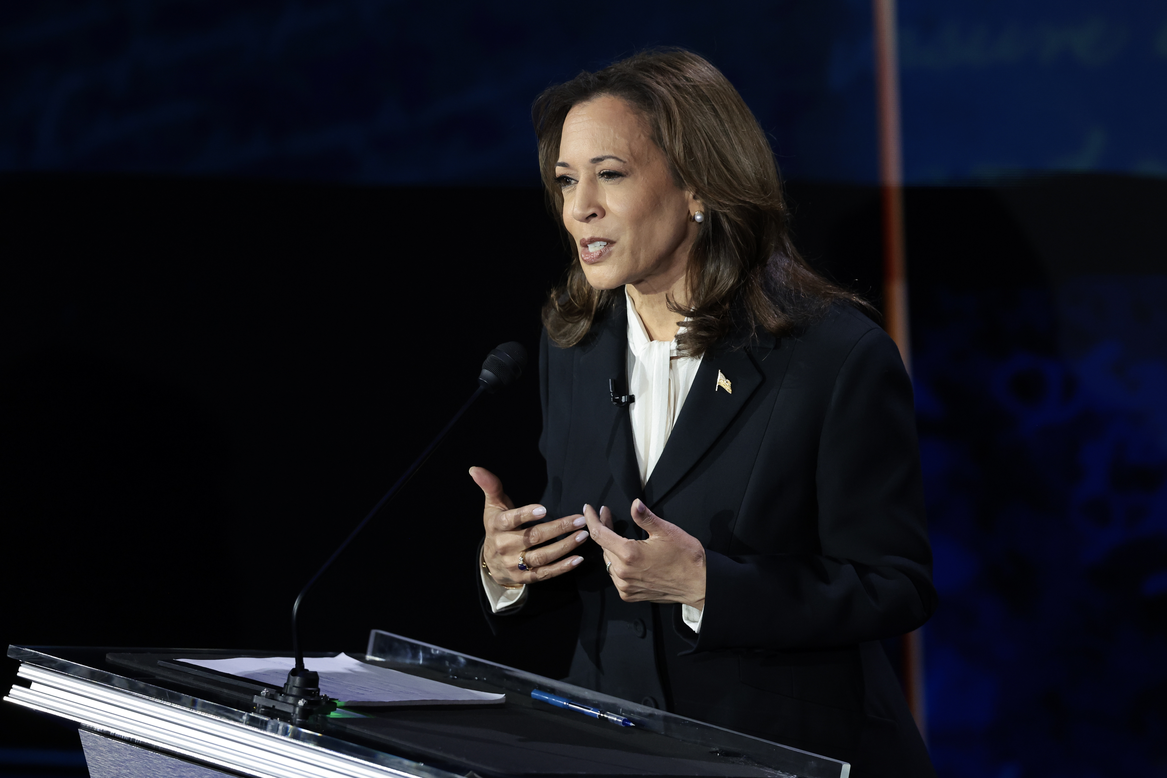 Kamala Harris stands at a podium, speaking into a microphone during an event. She is wearing a tailored suit with a small pin on her lapel