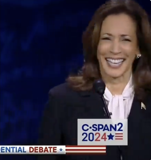 Kamala Harris is smiling at a podium during the C-SPAN2 Presidential Debate. She is wearing a dark blazer with a white blouse