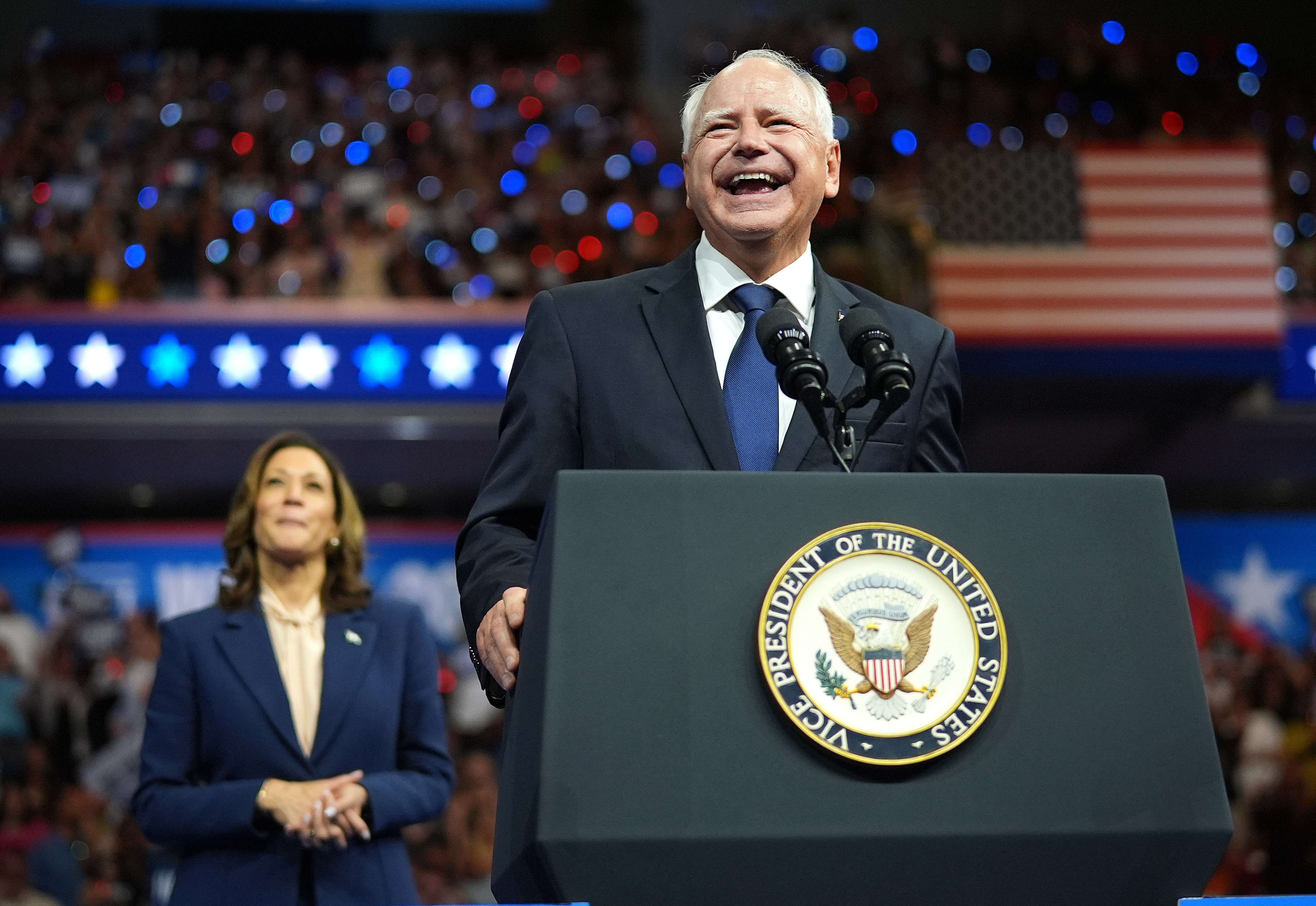 U.S. Vice President Kamala Harris stands behind Minnesota Governor Tim Walz, who is speaking and smiling at a large event with a crowd and American flags