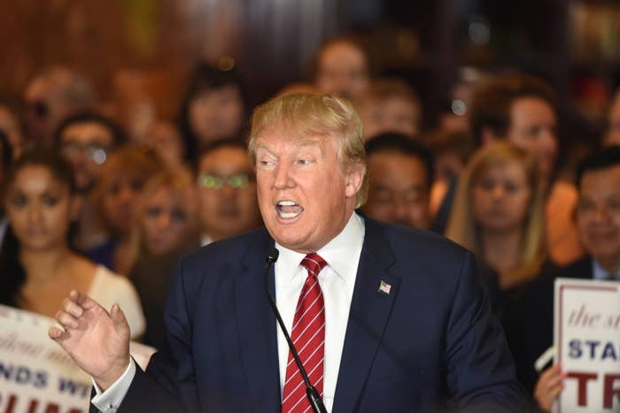 Donald Trump speaks to a crowded audience, some holding signs, at what appears to be a campaign event