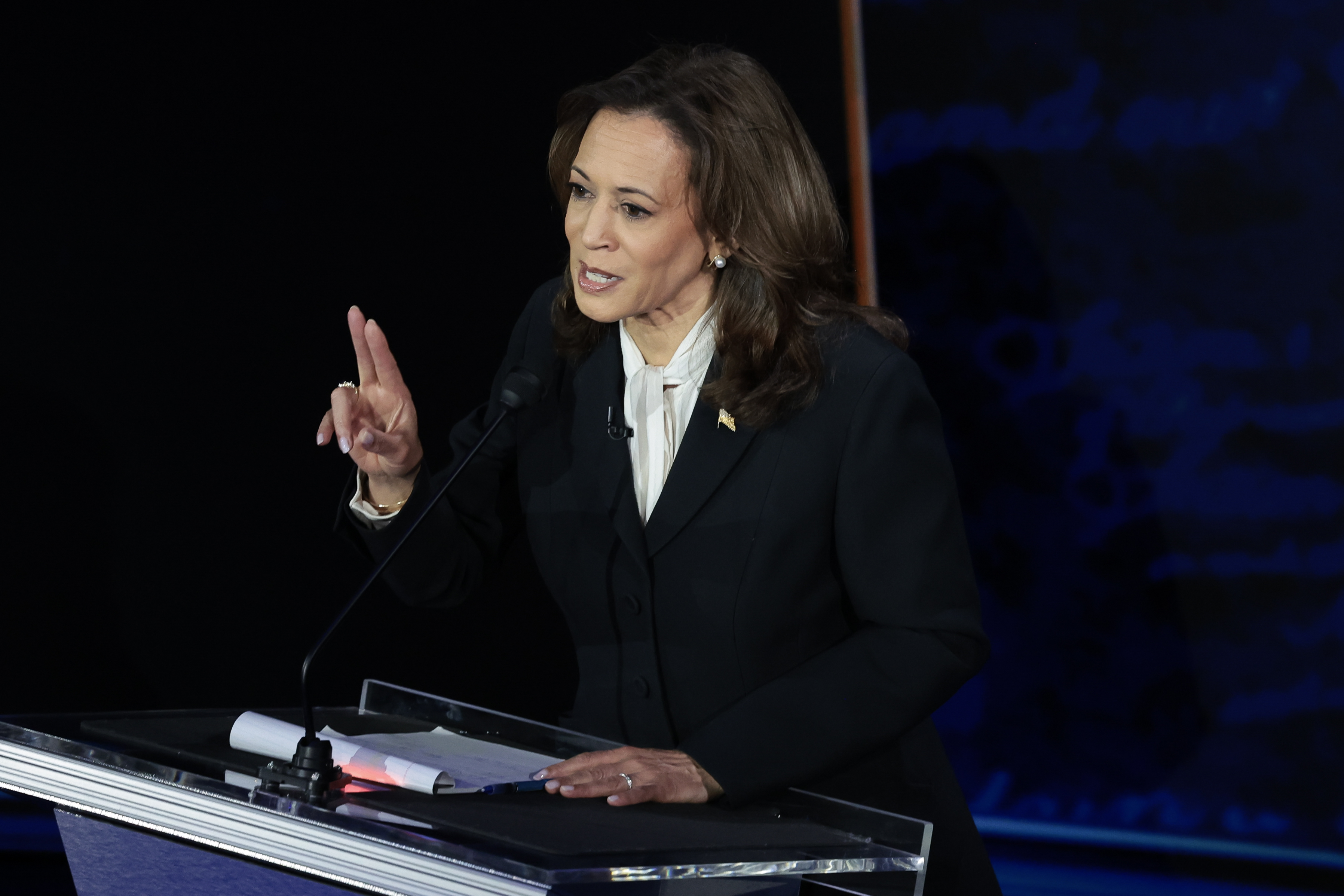 Kamala Harris stands at a podium, speaking, and gesturing with her right hand during a debate. She is wearing a business suit