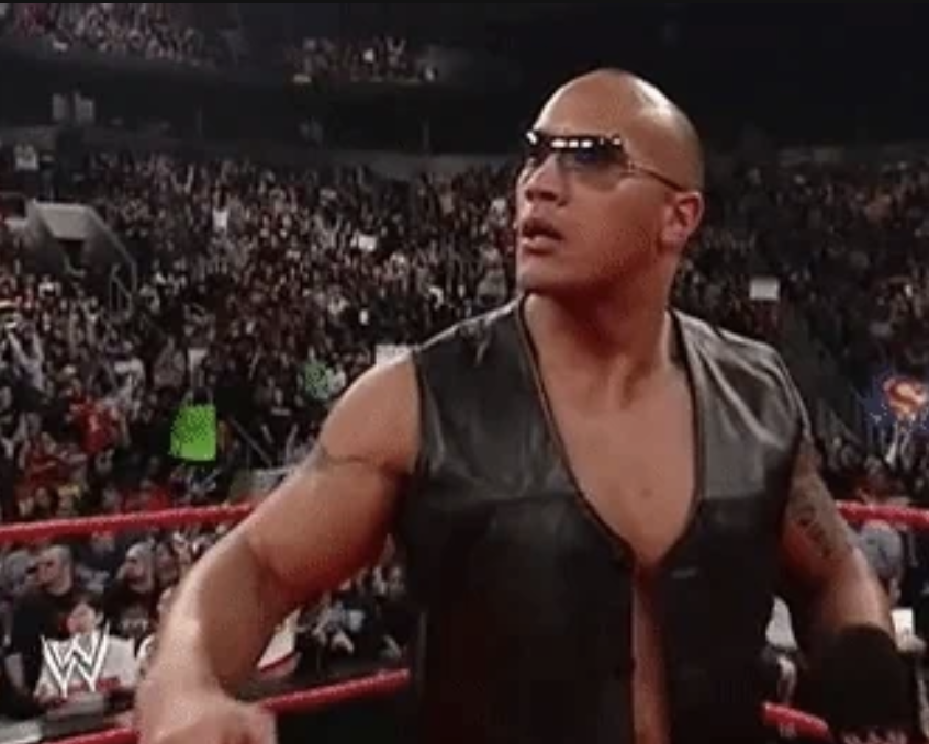Dwayne "The Rock" Johnson in wrestling ring wearing sunglasses and sleeveless black leather vest, with a crowd in the background