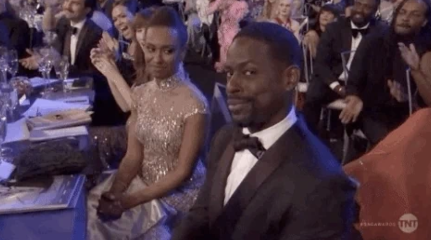 Sterling K. Brown at an event, seated and smiling at the camera, while people around him clap and smile. Another person is seated beside him