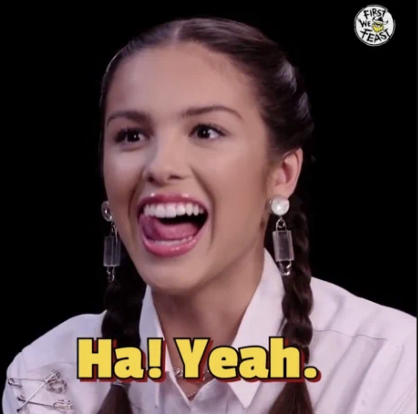 Olivia Rodrigo sticks her tongue out and smiles, wearing braided hair and a white shirt with silver safety pin details. "Ha! Yeah." text is displayed
