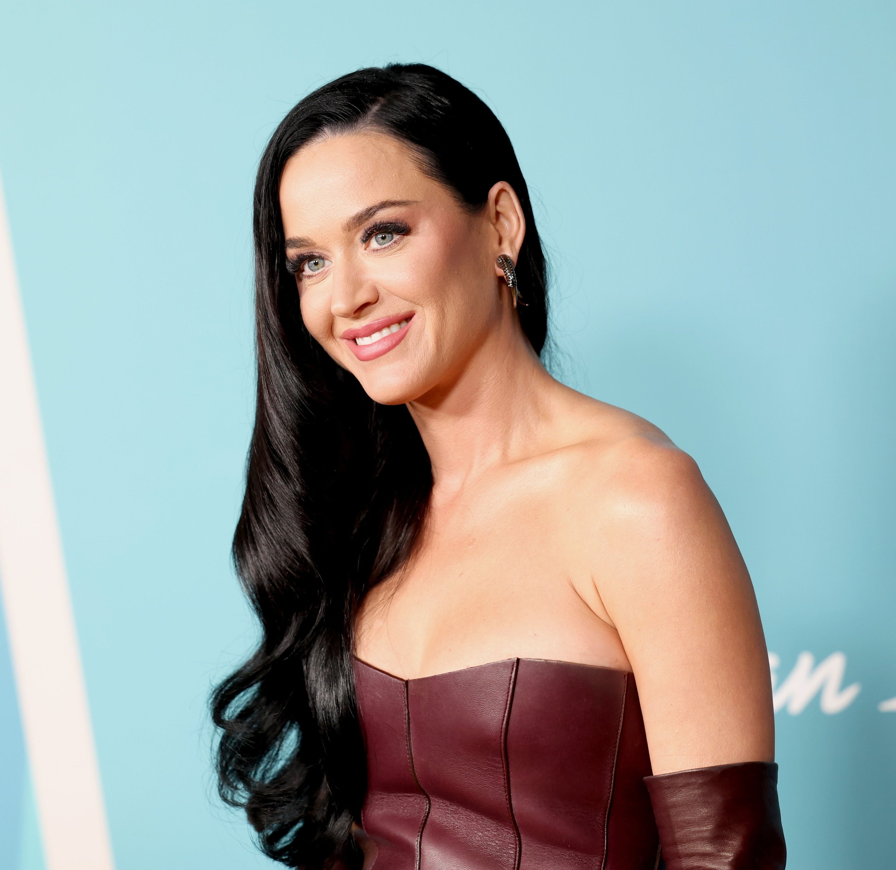 Katy Perry at an event, wearing a strapless maroon leather dress with long matching gloves, smiling and looking to the side