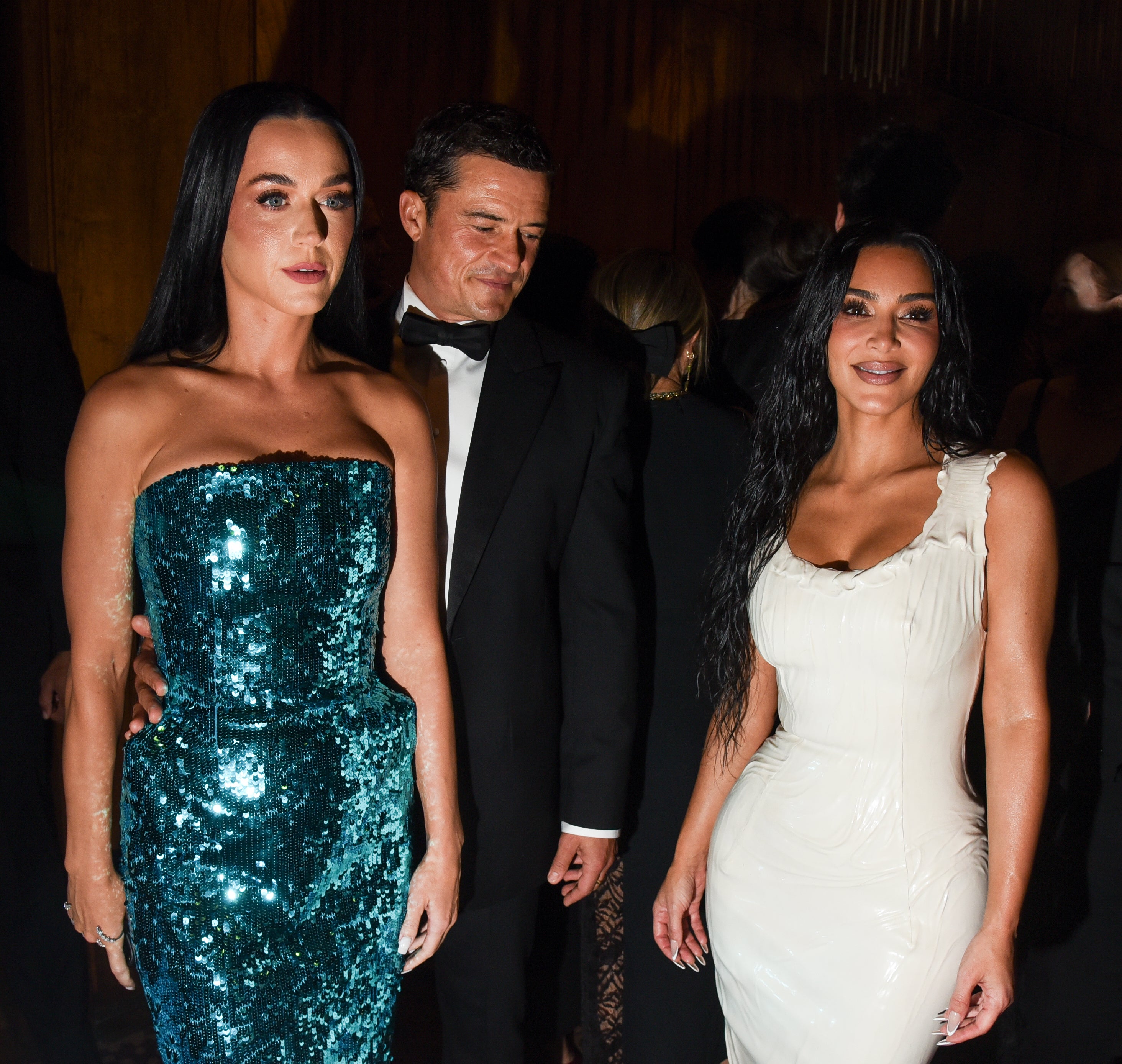 Katy Perry in a sparkling strapless gown and Kim Kardashian in a sleek fitted dress attend an event with several other people in the background