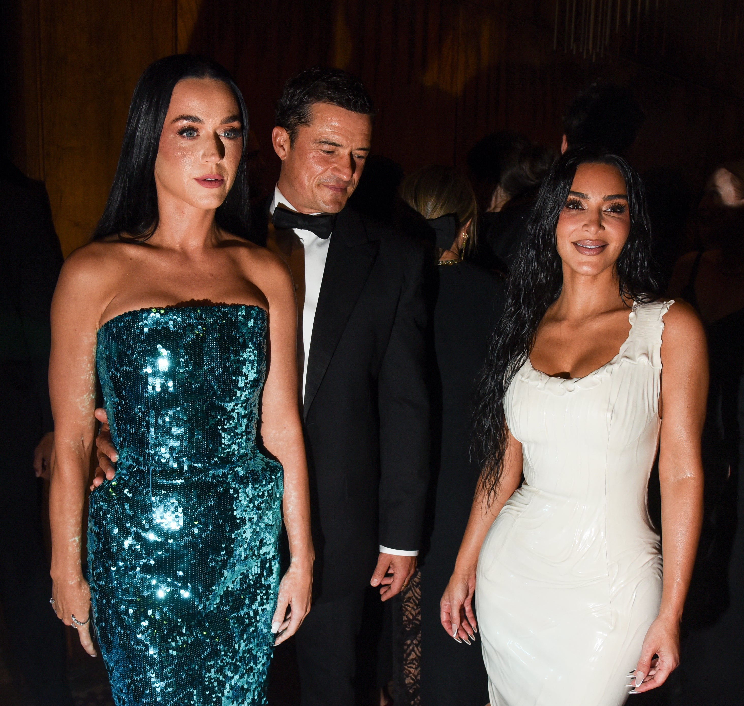 Katy Perry in a sparkling strapless gown and Kim Kardashian in a sleek fitted dress attend an event with several other people in the background