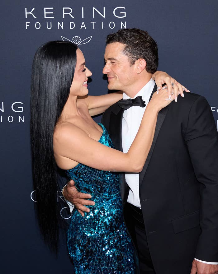 Katy Perry and Orlando Bloom embrace and smile at each other on the red carpet at a Kering Foundation event. Katy wears a sequined dress, and Orlando wears a tuxedo