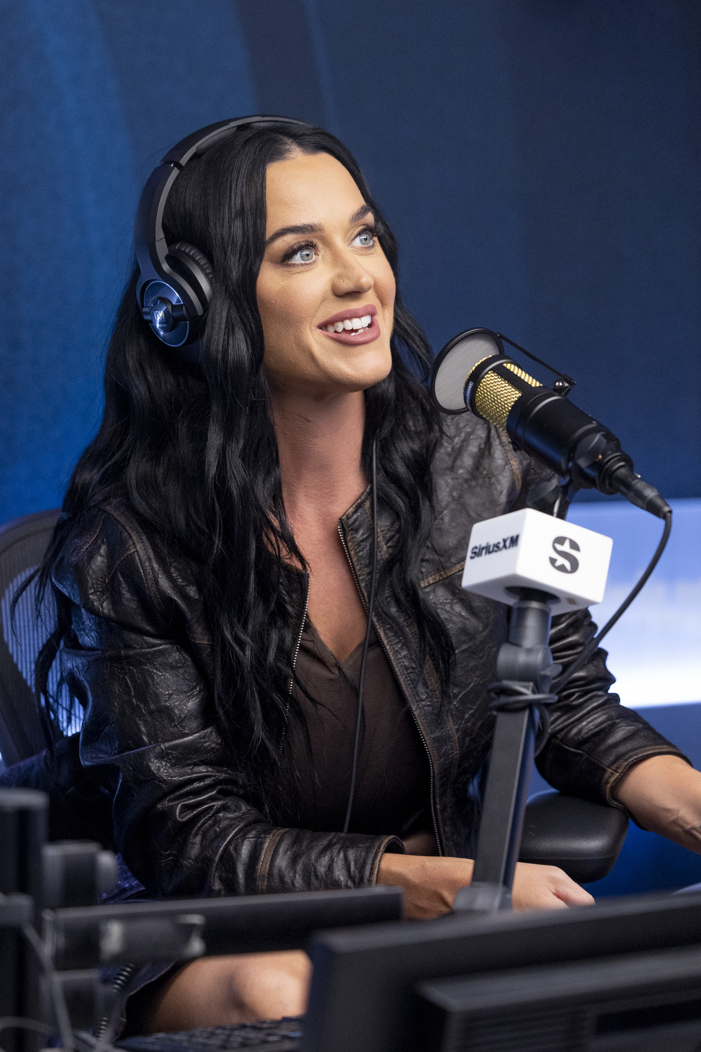 Katy Perry is seated in a radio studio wearing headphones, speaking into a microphone. She is dressed in a casual outfit with a leather jacket