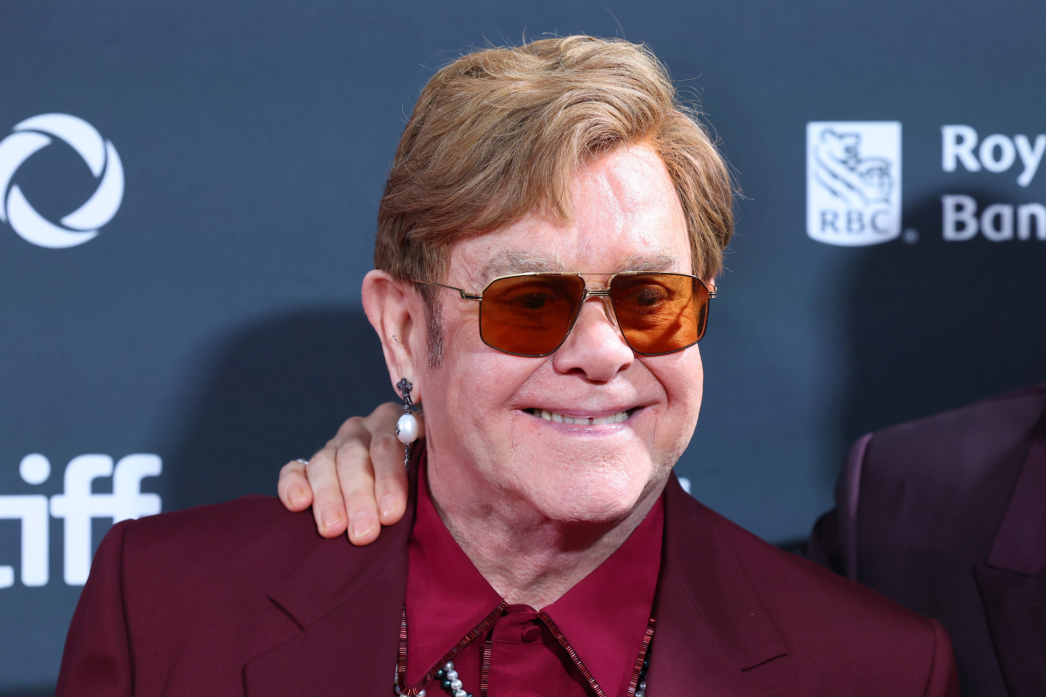 Elton John wears a maroon suit and orange-tinted glasses, smiling at a public event. An unidentified person has their hand on his shoulder in the background