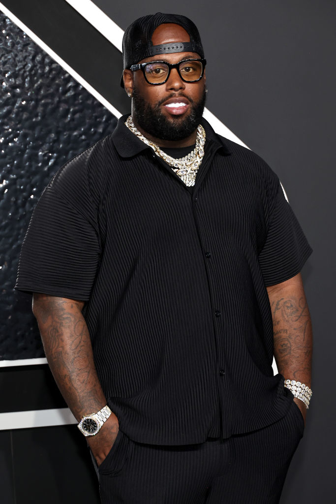 Demarcus Stigar is posing on a red carpet wearing glasses, a baseball cap, a pinstripe short-sleeve shirt, matching pants, and multiple silver chains and bracelets