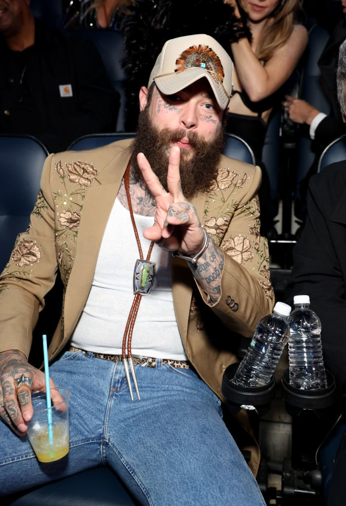 Post Malone flashes peace sign, wearing a beaded jacket, t-shirt, jeans, and a feathered cap sitting at an event