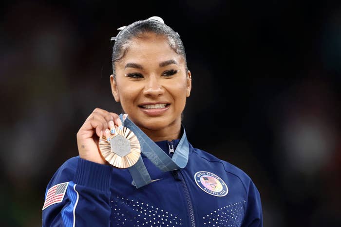 Olympian gymnast Jordan Chiles smiles, holding her bronze medal. She wears a team USA tracksuit with an American flag patch on the sleeve