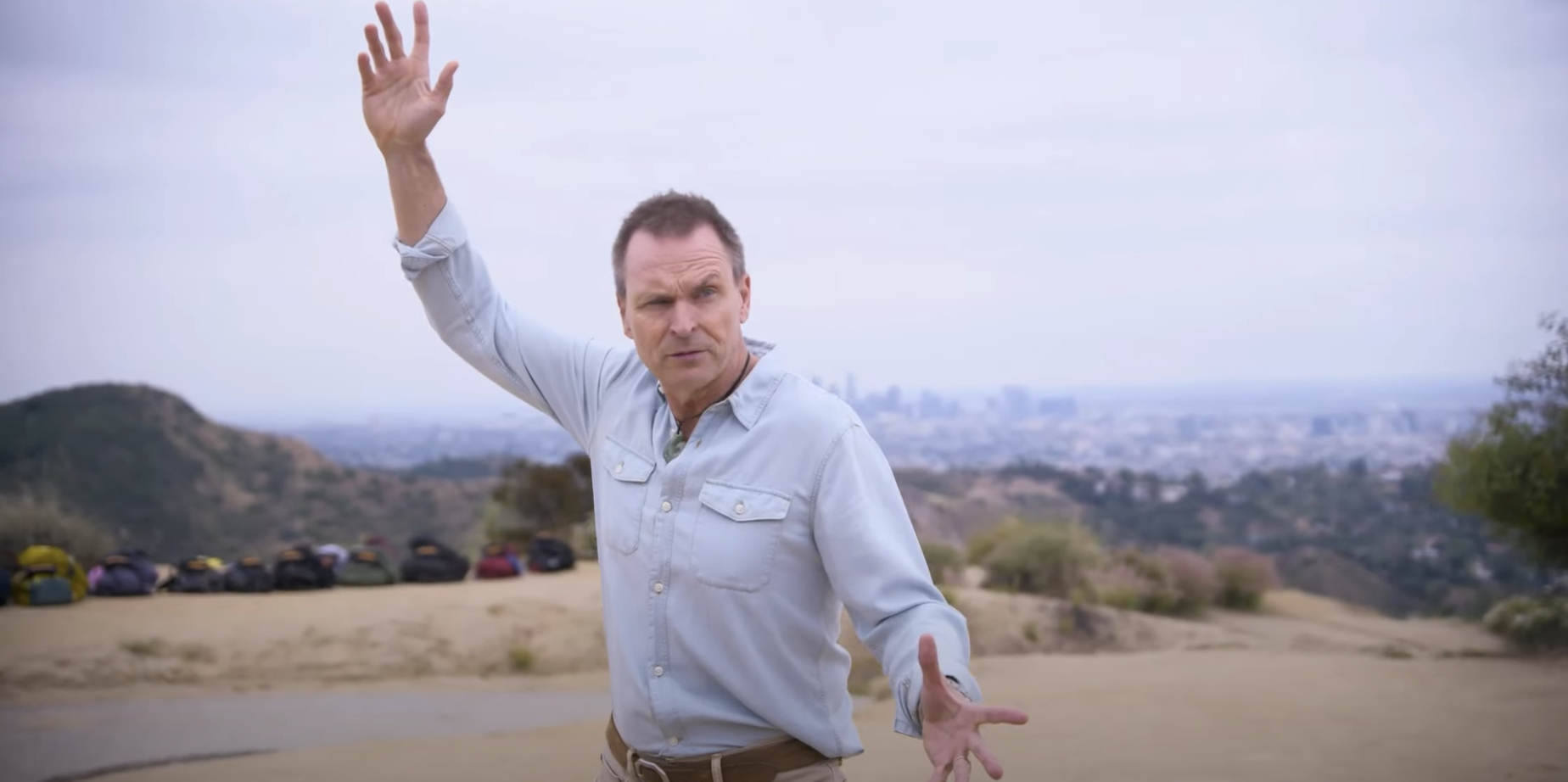 Phil Keoghan stands outdoors, raising his hand, with a cityscape in the background