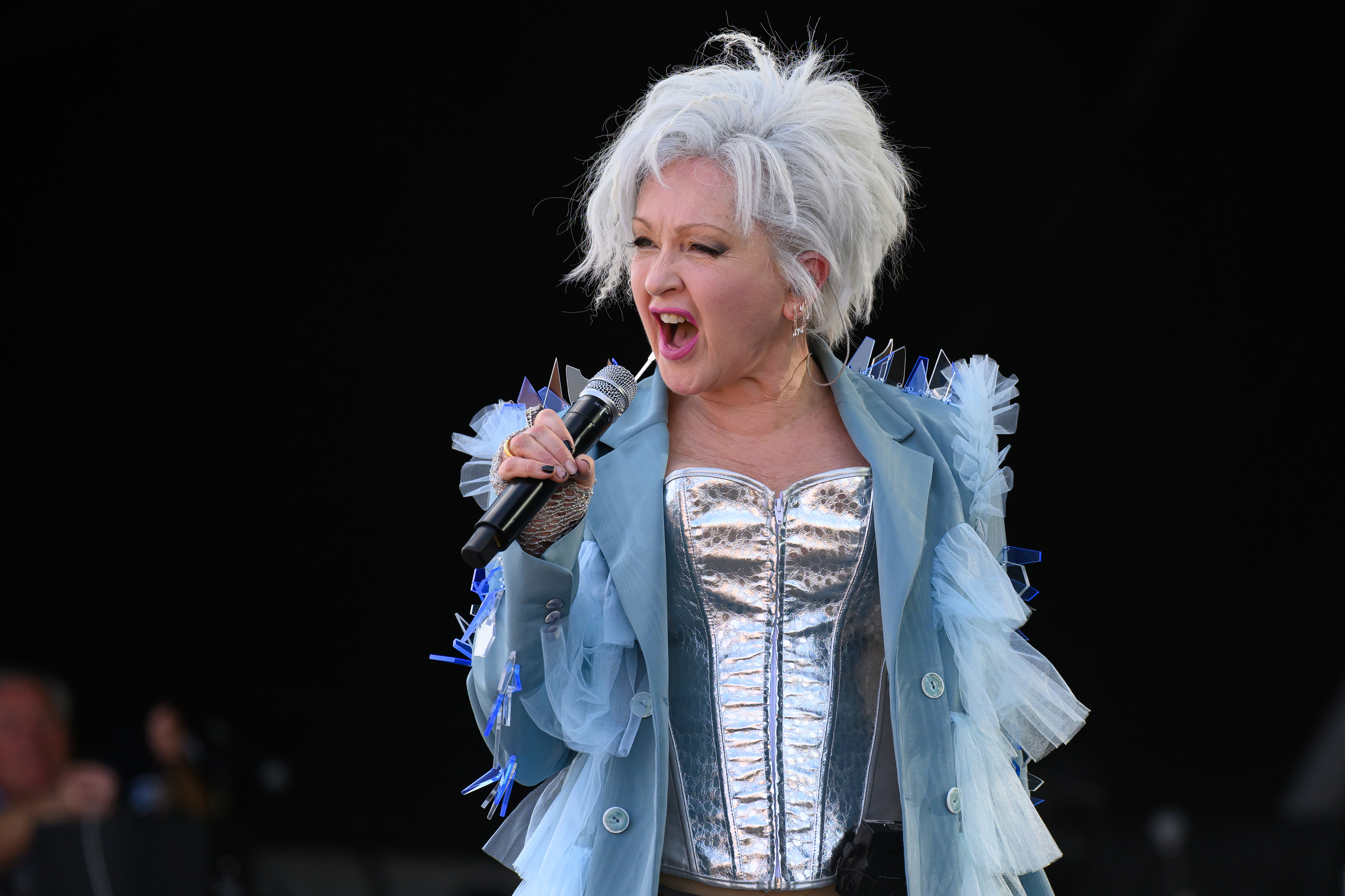 Cyndi Lauper performing on stage wearing a metallic corset and a sheer, ruffled jacket. She holds a microphone while passionately singing