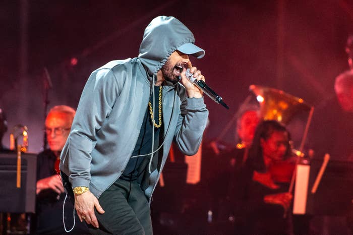 Eminem performs on stage wearing a hoodie and cap, holding a microphone. Musicians with instruments can be seen in the background