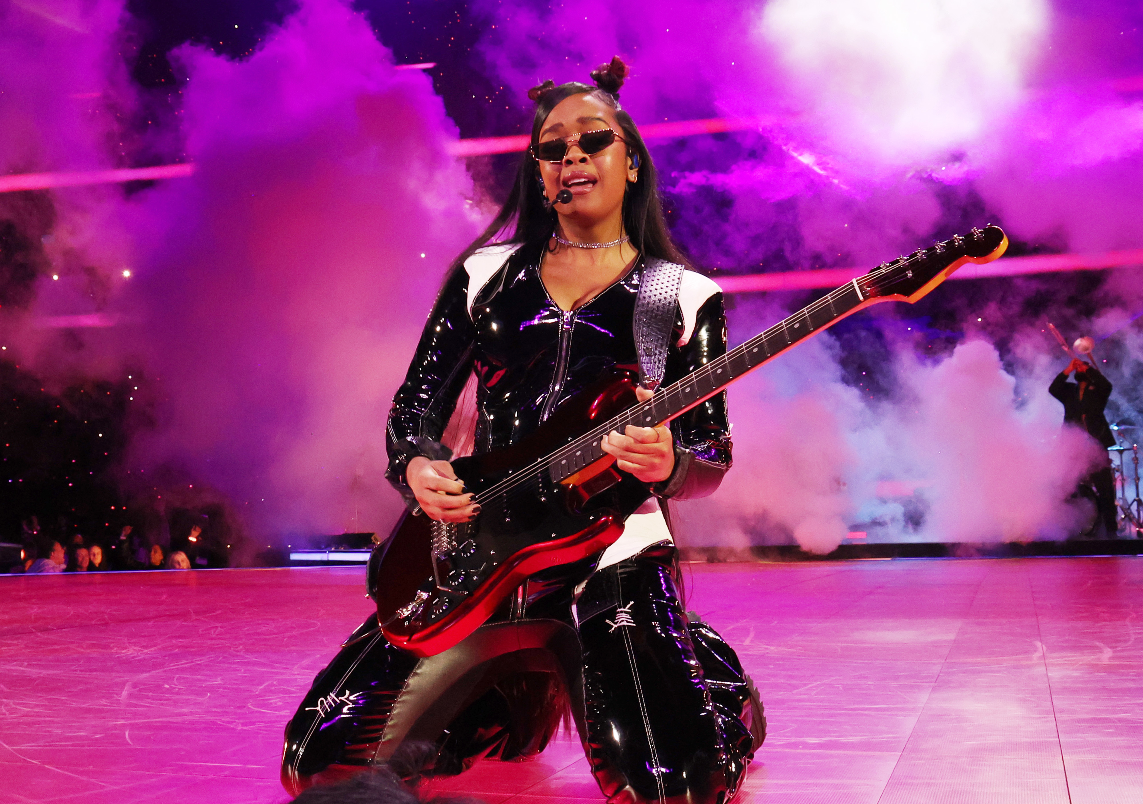H.E.R. performs on stage wearing a black leather outfit with a guitar, surrounded by smoke effects