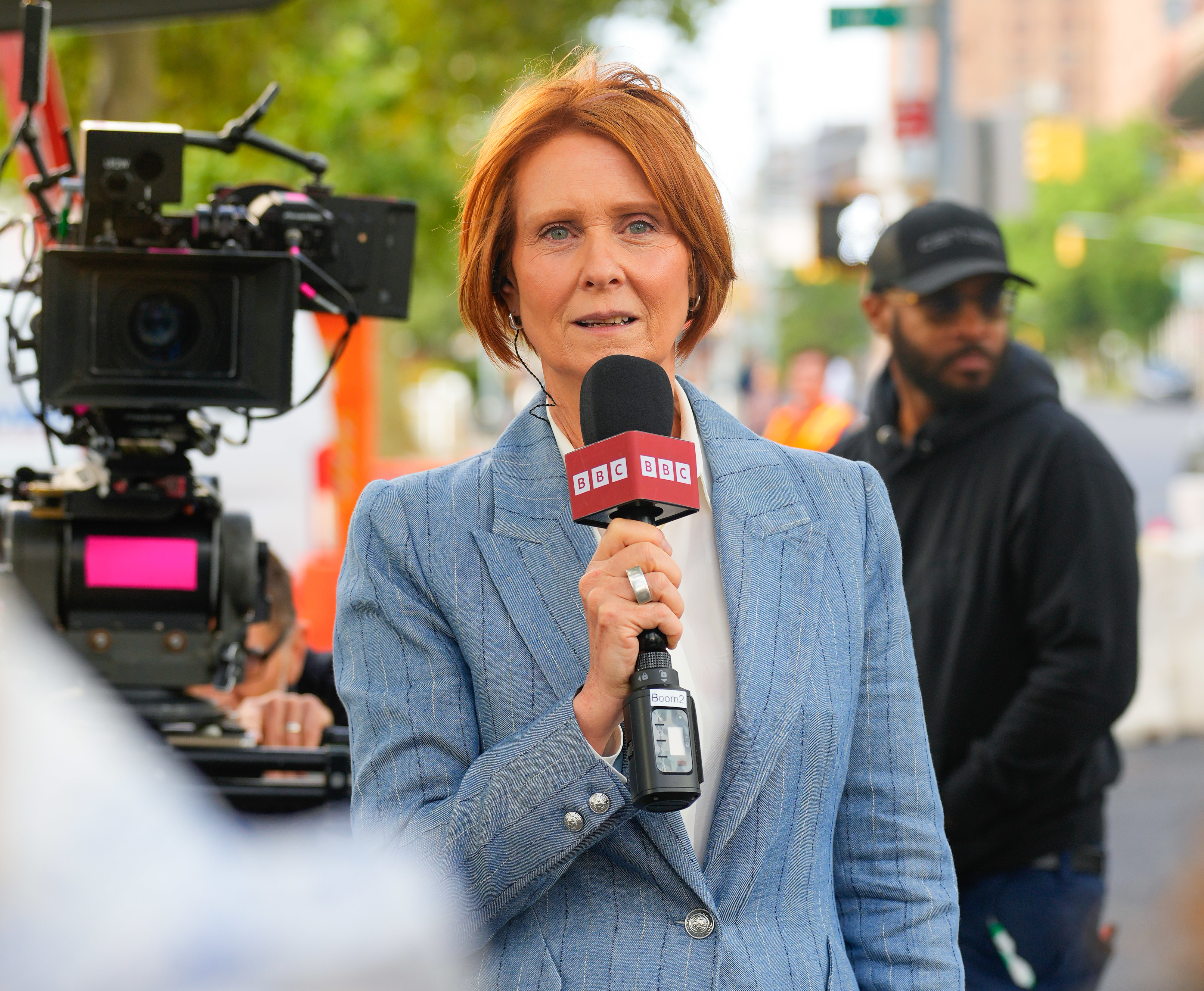 Cynthia Nixon stands holding a BBC microphone, in a light-colored blazer, while filming outdoors with a cameraman and crew in the background