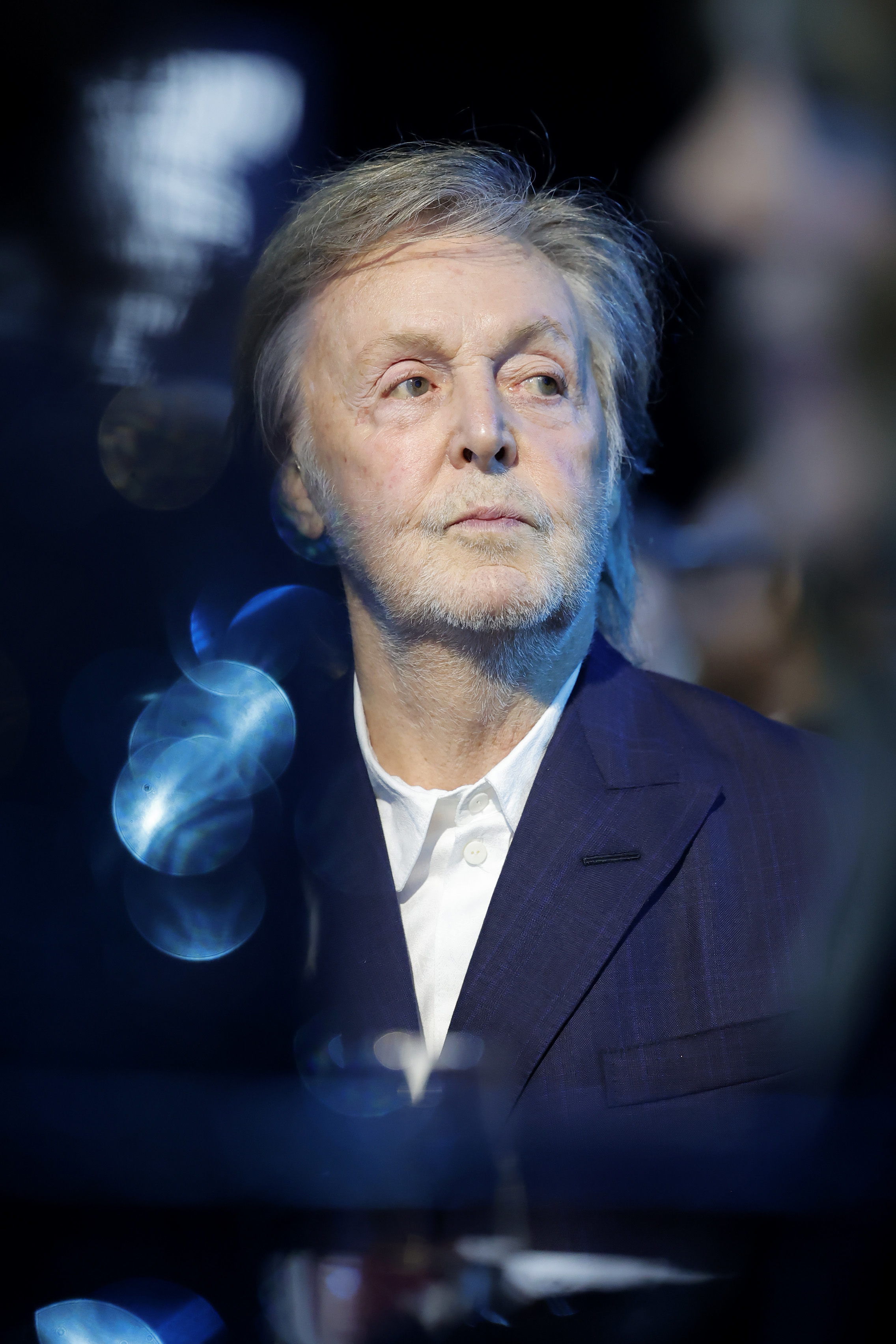 Paul McCartney wearing a formal suit, looking thoughtful, in a candid moment at an event