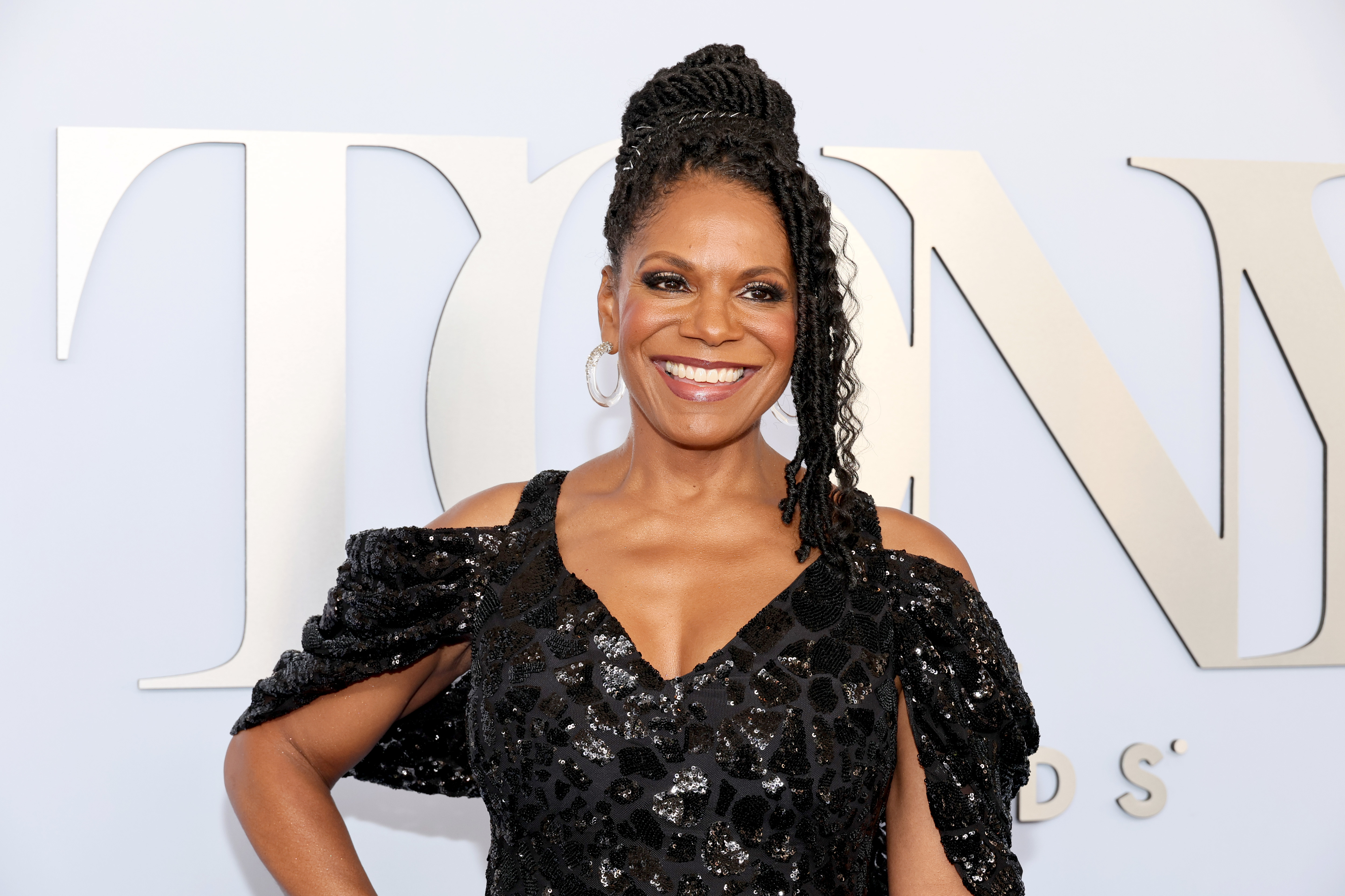 Audra McDonald smiles on the red carpet at the Tony Awards, wearing a sparkling black gown with open shoulders and braided updo hairstyle