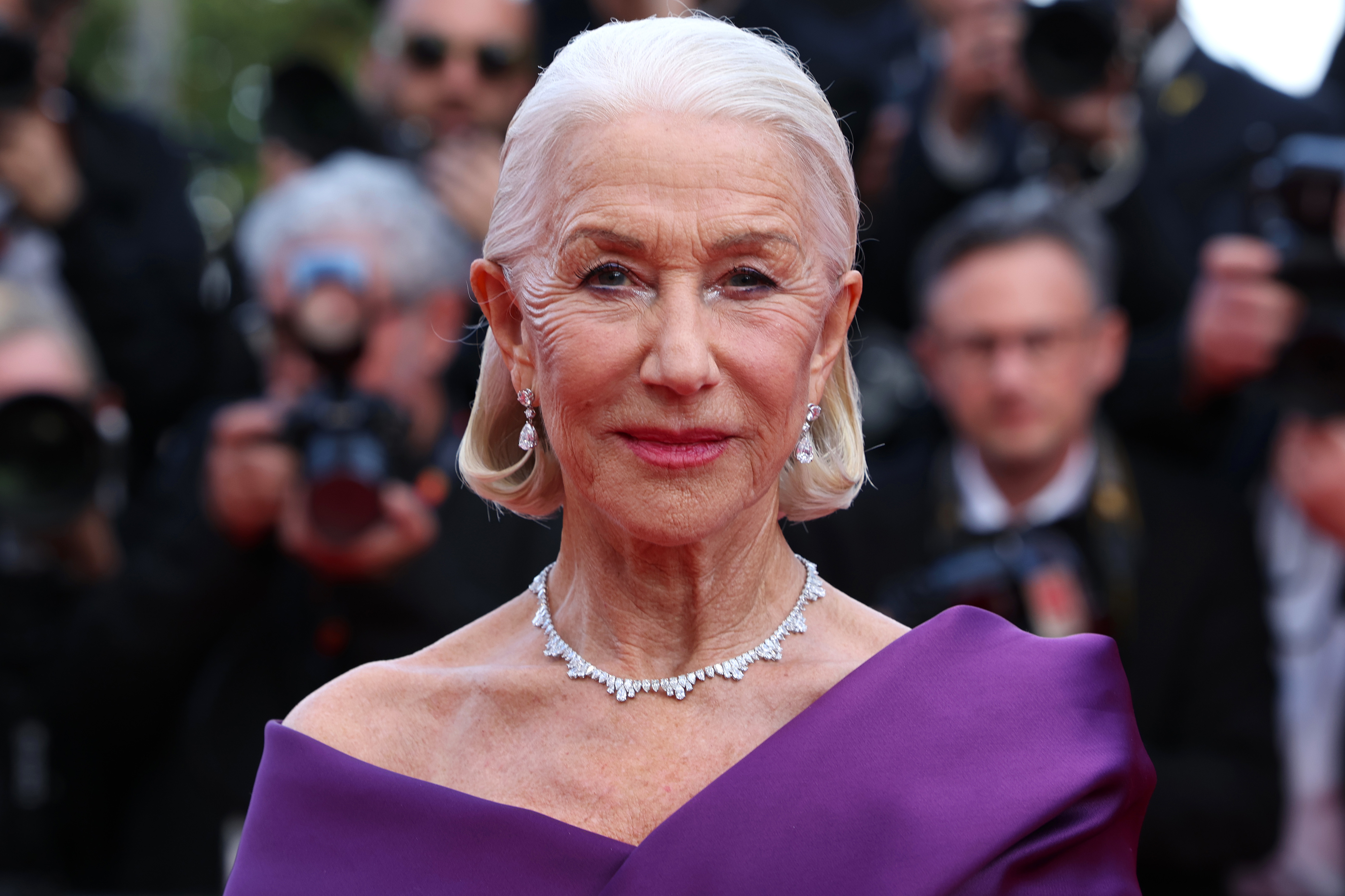 Helen Mirren on the red carpet, wearing an elegant off-the-shoulder gown and diamond jewelry, with photographers and onlookers in the background