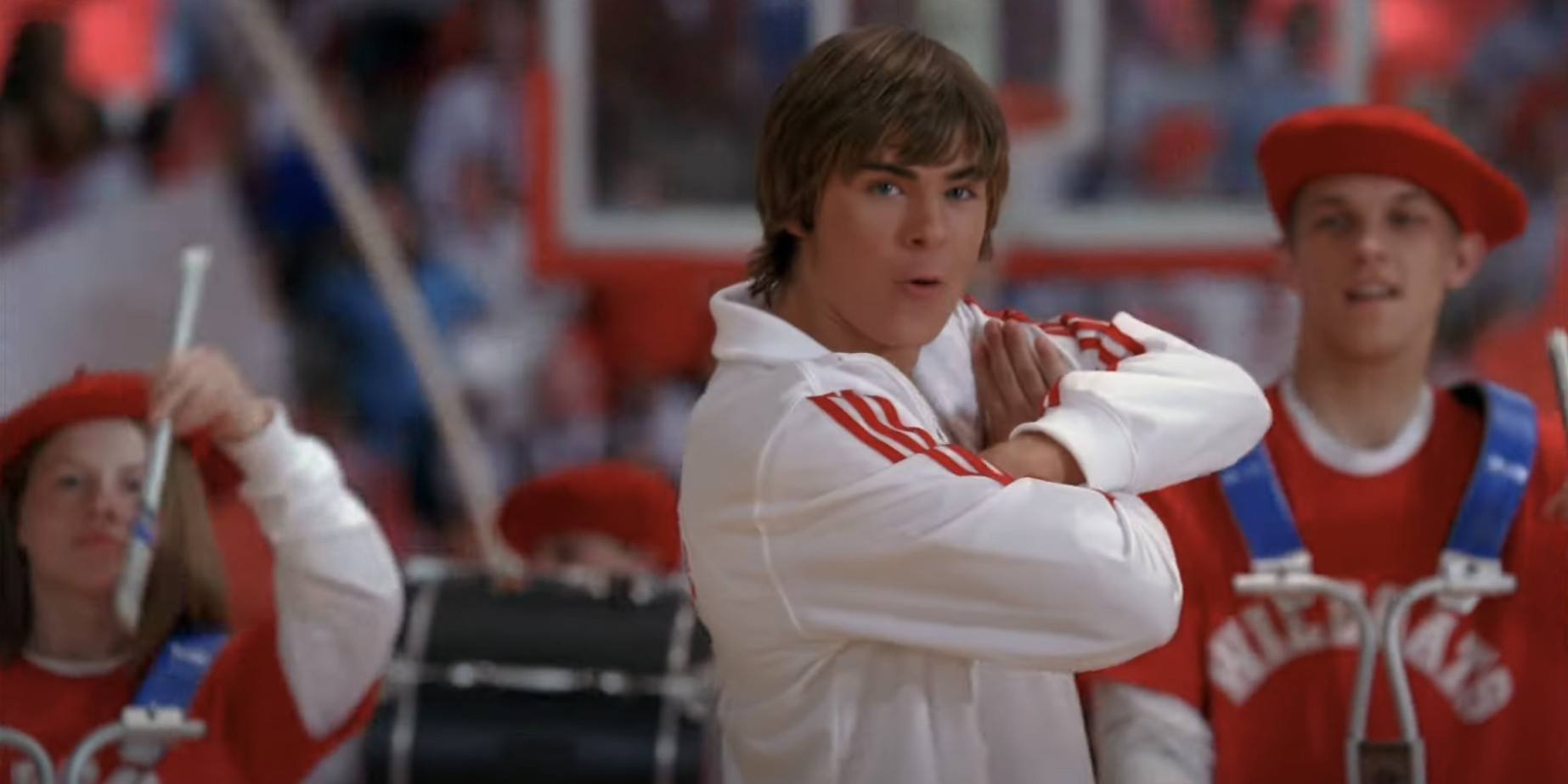 Zac Efron performs a dance routine in a gymnasium, wearing a white jacket with red stripes, surrounded by drummers in red and white costumes