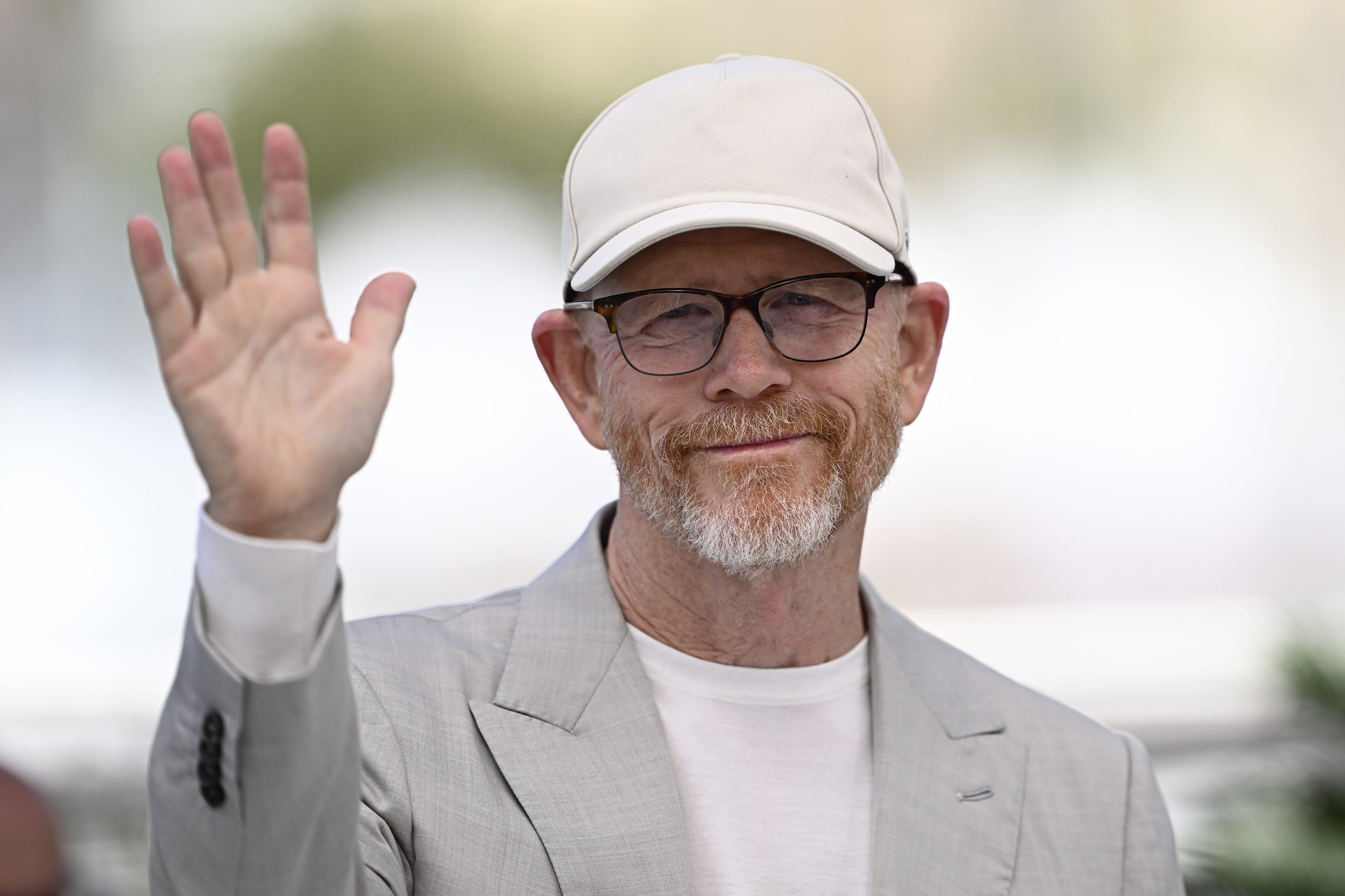 Ron Howard waves while wearing a casual light-colored cap, glasses, a light suit jacket, and a white shirt