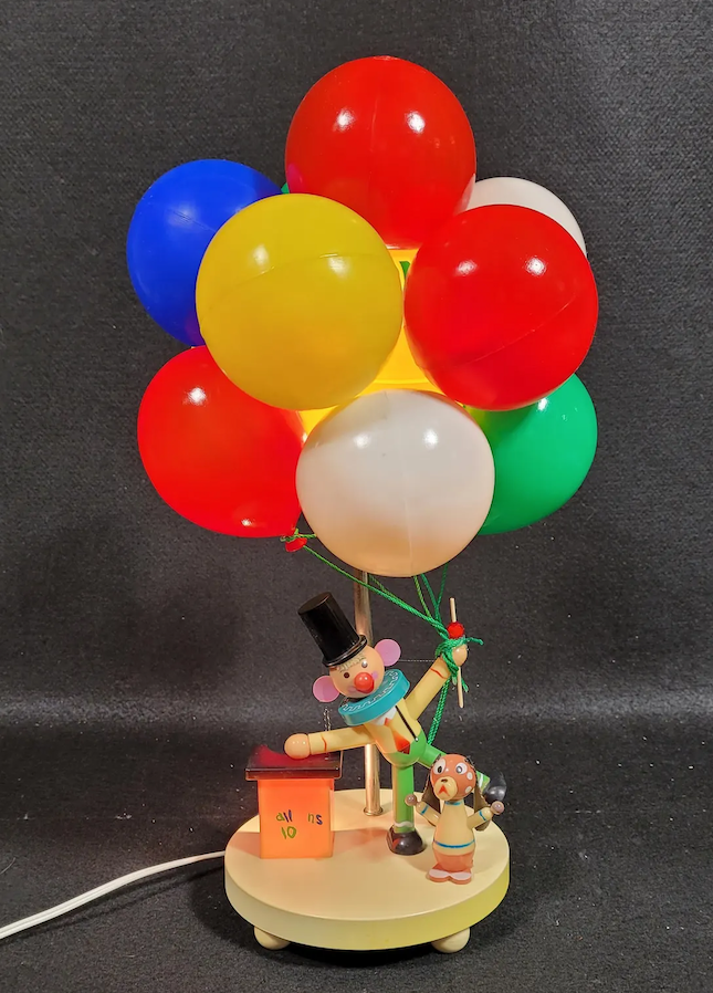 A lamp featuring a clown holding a bunch of colorful balloons, with a small dog figure and a red box labeled with "all is" near the base