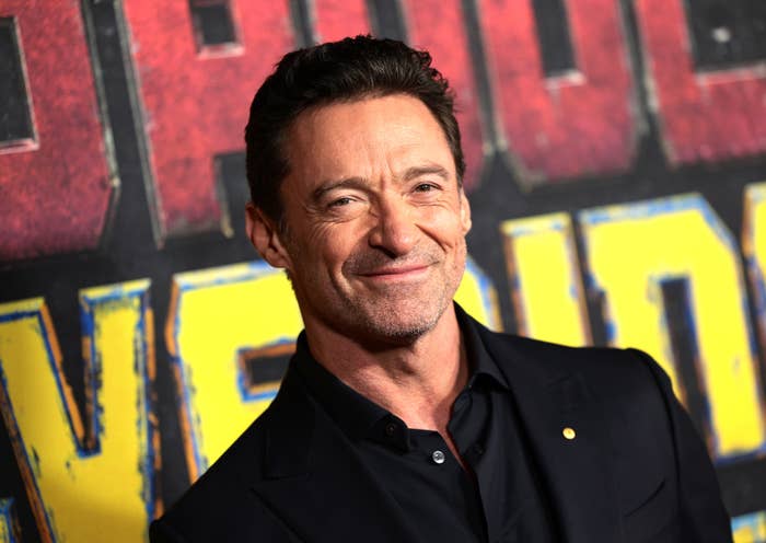 Hugh Jackman smiles at a movie premiere, wearing a black suit and black shirt