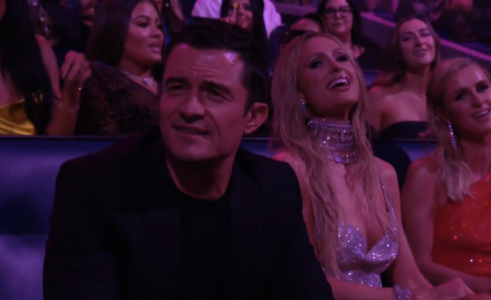 Orlando Bloom, Paris Hilton, and two others sit among an audience. Paris Hilton wears a sparkling dress with a choker necklace