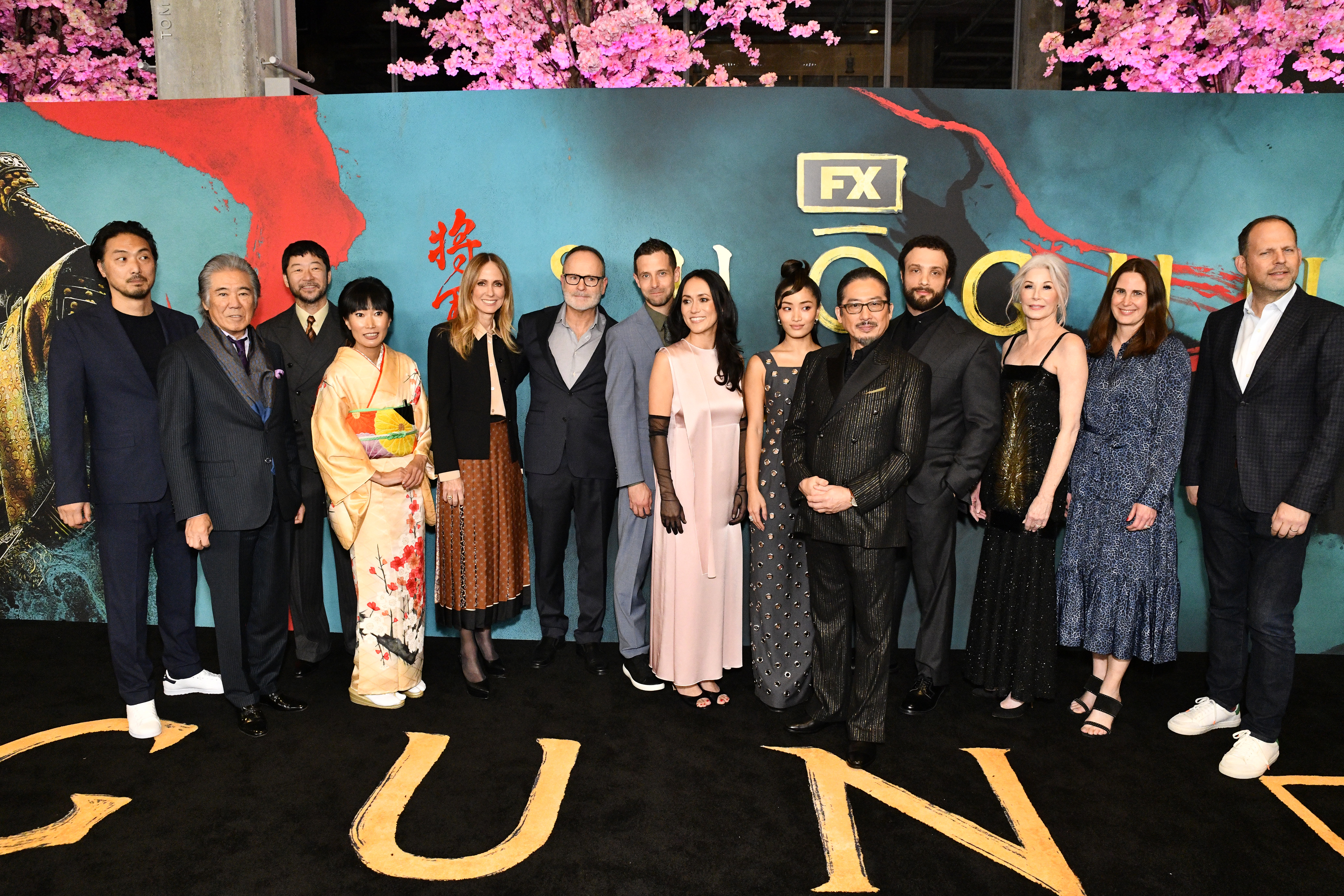 Group of actors and creators from FX's "Shogun" TV series stand on black carpet at the premiere, dressed in formal and stylish attire including suits and dresses