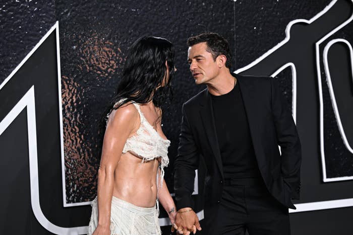 Katy Perry in a midriff-baring semi-sheer, fringed outfit, holds hands with Orlando Bloom in a dark suit as they interact on a red carpet with a textured backdrop