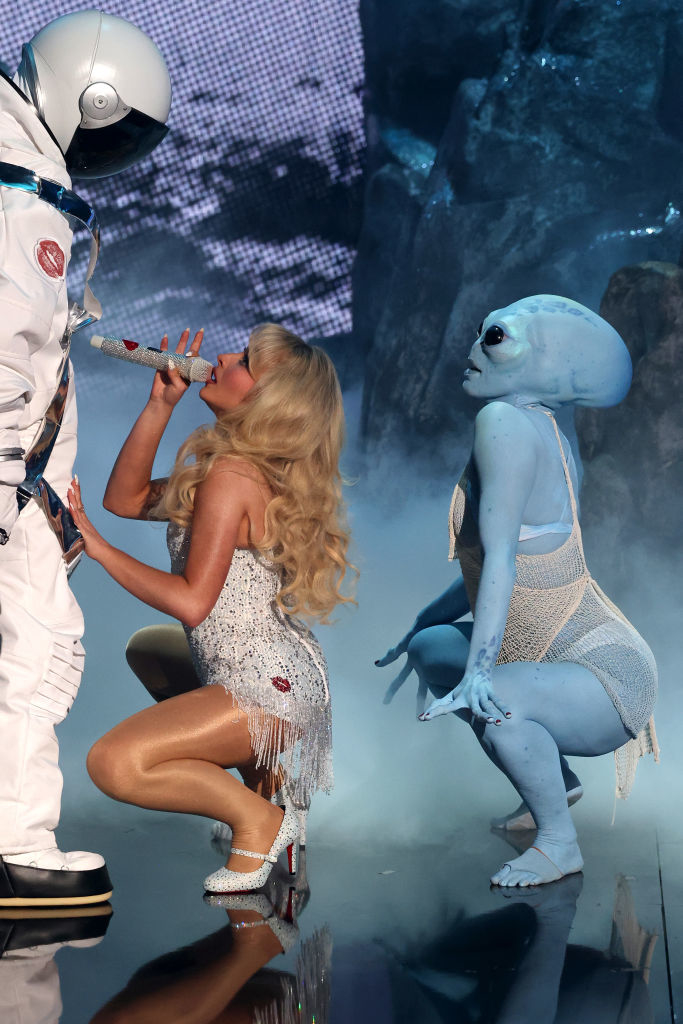 Sabrina Carpenter, dressed in a sparkling outfit with high heels, squatting in front of a person in an astronaut suit and another person dressed as an alien squatting behind her