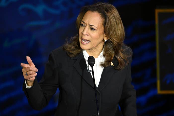 Kamala Harris speaks passionately on stage, gesturing with her hand, wearing a black suit with a white shirt