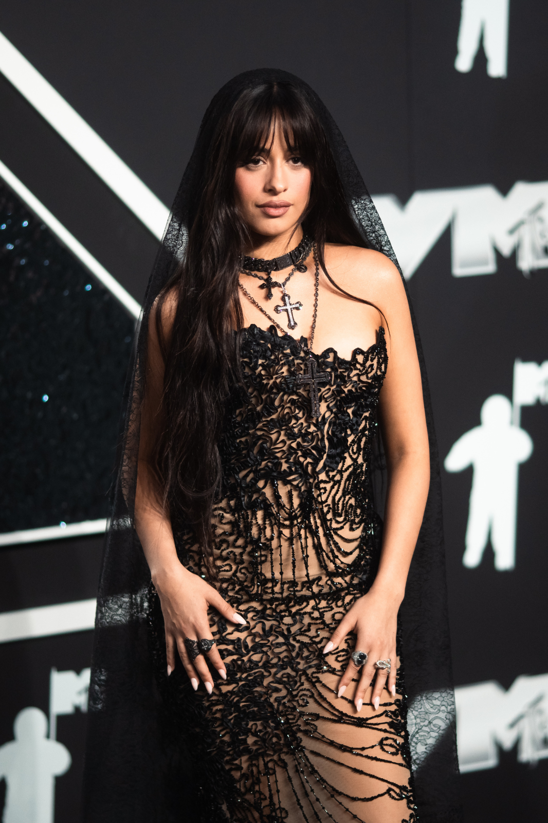 Camila Cabello lleva un vestido transparente con detalles de encaje y un velo negro en un evento de alfombra roja.