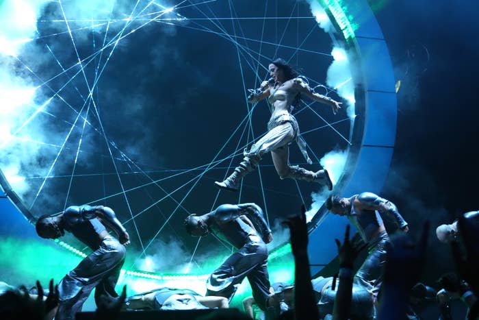 Five dancers perform a routine on stage with futuristic props and special effects while Katy Perry, dressed in a sci-fi-inspired outfit, floats above them