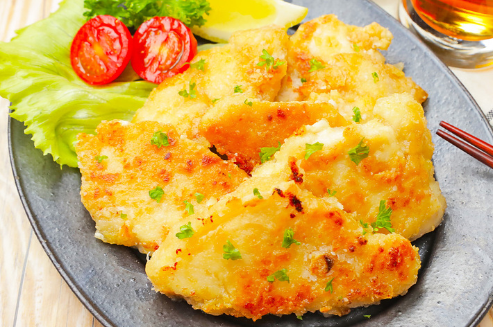 レタスとトマトスライスが添えられた白身魚のフライが黒い皿に盛られた料理。日本風の盛り付け。