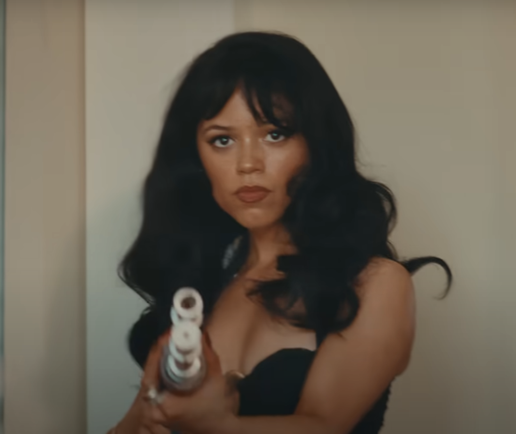 Jenna Ortega in the Taste music video, wearing a strapless top, holds a prop shotgun while looking intently at the camera