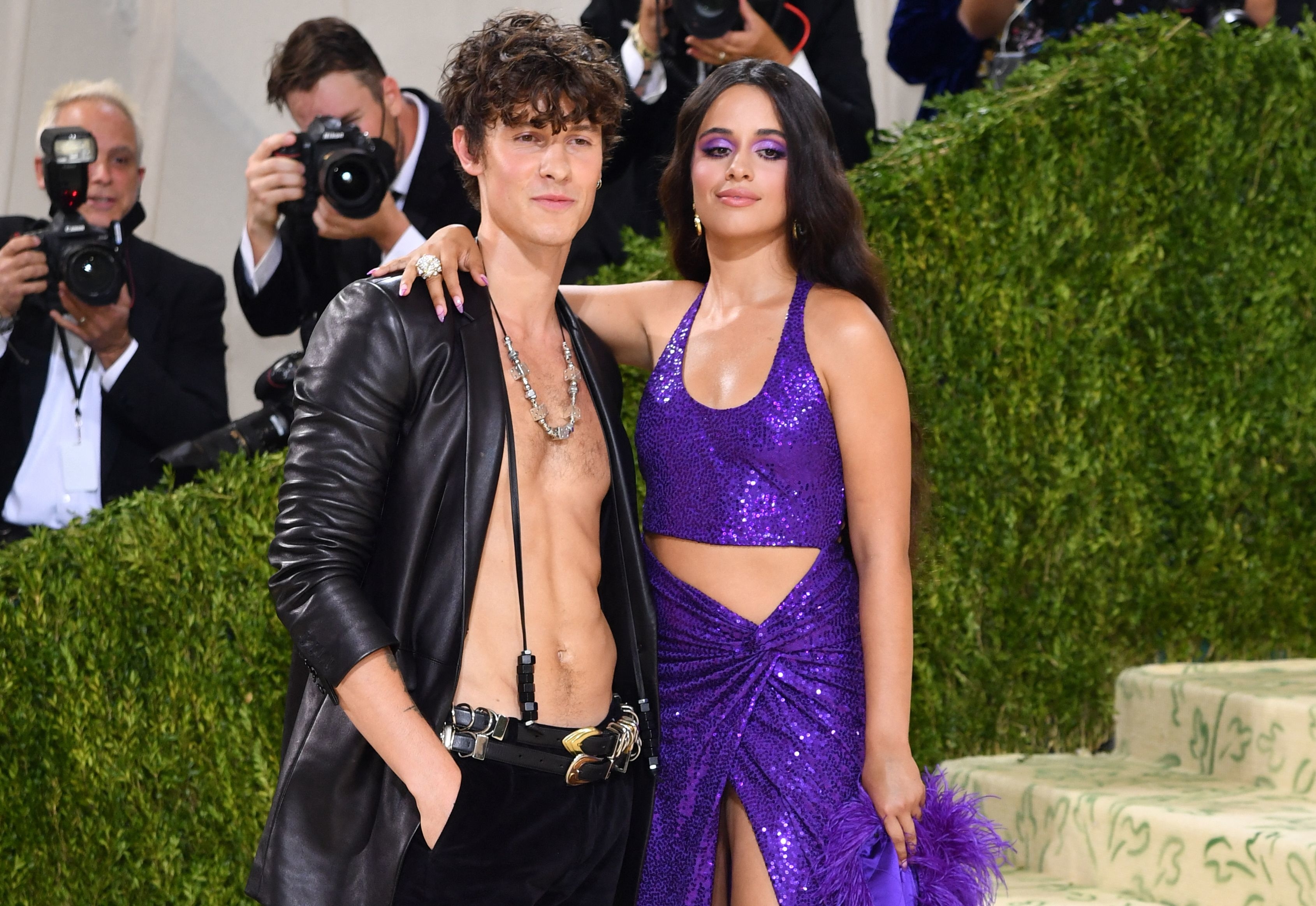 Shawn Mendes, in an open leather jacket and dark pants, and Camila Cabello, in a sparkling dress, pose on the Met Gala red carpet. Photographers in the background