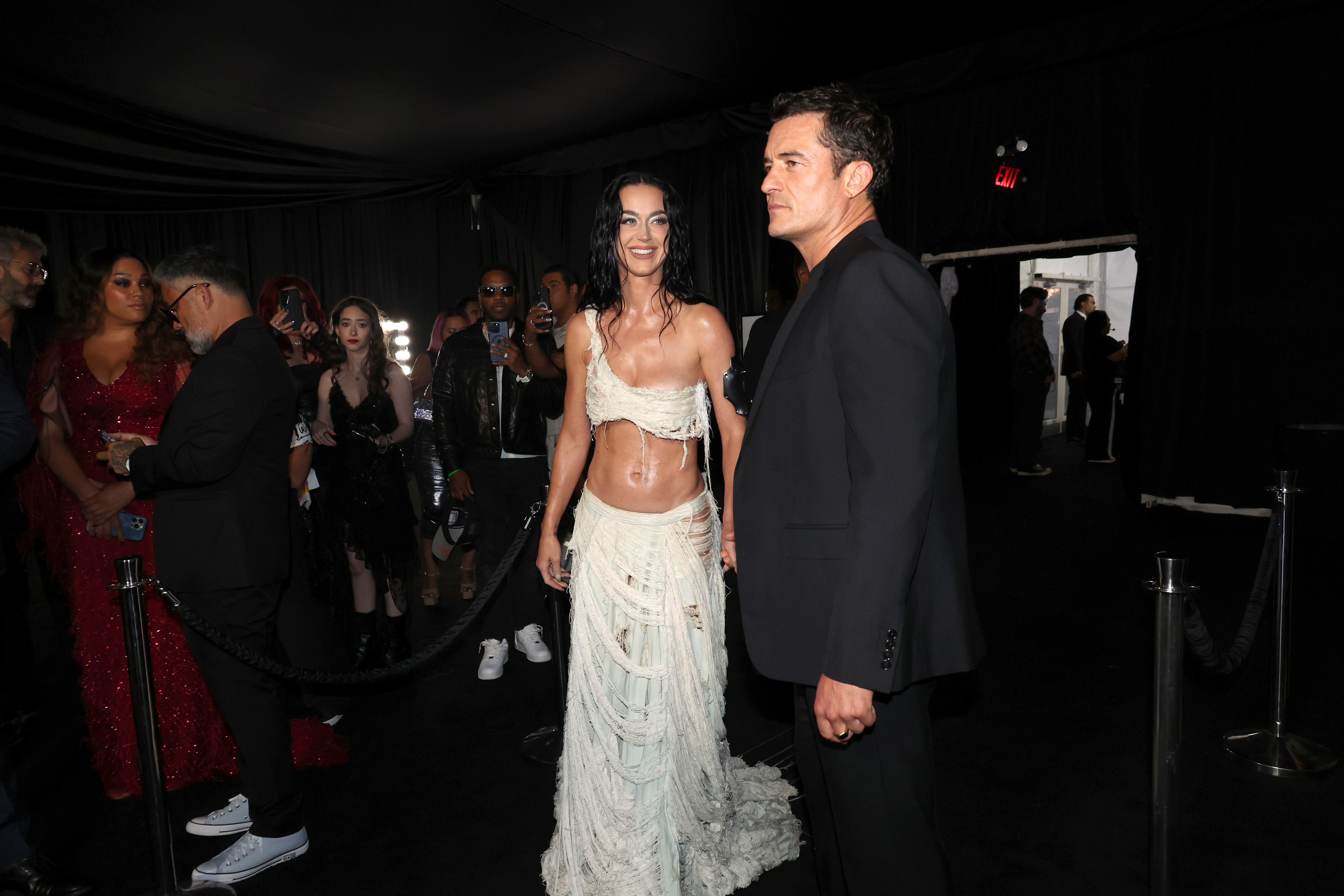 Katy Perry in a white, fringed outfit and Orlando Bloom in a dark suit at an event, with a crowd in the background