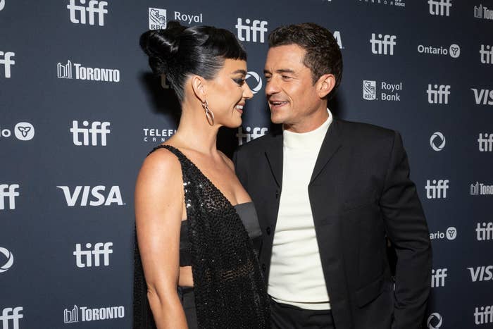 Katy Perry in a dark sequined dress and Orlando Bloom in a dark suit smile at each other on the red carpet at TIFF