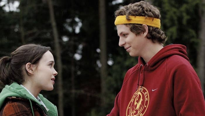 Elliot Page in a green vest and Michael Cera in a red hoodie with a yellow headband smile at each other outdoors