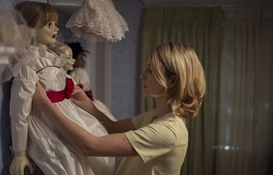 A woman arranges a doll wearing a white dress with a red band in a room with other dolls and a green curtain