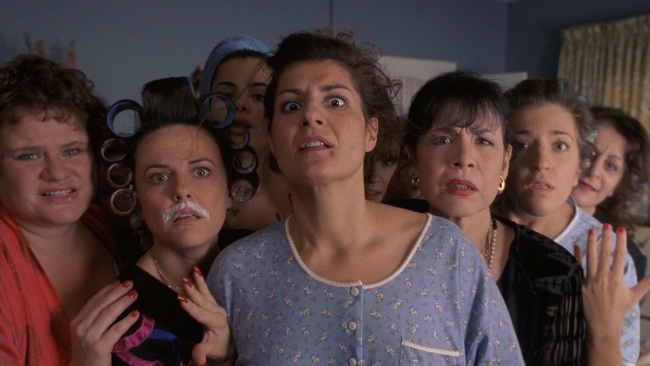 A group of women, including Nia Vardalos, stands closely together, each with a surprised or focused expression. Some wear hair rollers and facial cream